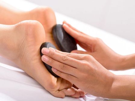 A person is receiving a hot stone foot massage with smooth black stones being pressed against the soles of their feet.