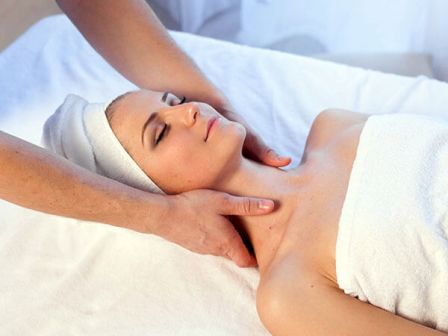 A person relaxing on a massage table with eyes closed, receiving a neck massage. The individual is wrapped in a white towel.