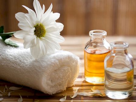 A rolled white towel, a white flower, and two small bottles—one with an amber liquid and one with a clear liquid—are arranged on a wooden surface.