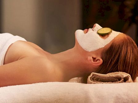 A person is lying down with a white facial mask and cucumber slices on their eyes, relaxing on a towel. The scene looks like a spa setting.