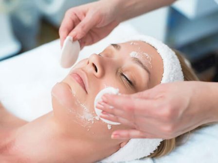 A person is receiving a facial treatment with a headband on, while someone applies a cream or mask to their face using cotton pads.