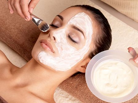 A person is receiving a facial treatment, with a white mask being applied using a brush while holding a bowl of the same substance.