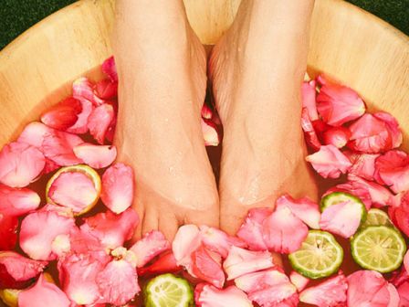Two feet are soaking in a wooden basin filled with water, pink rose petals, and slices of green lime.