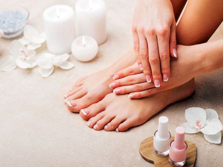 A person's manicured hands rest on their pedicure feet, surrounded by candles, flowers, and nail polish bottles, suggesting a spa setting.