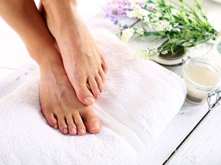 The image shows a pair of well-groomed feet resting on a white towel, surrounded by flowers and spa products, suggesting a relaxing spa treatment.