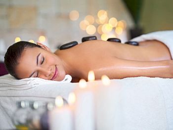 A woman is receiving a hot stone massage while lying down, with lit candles and a relaxed ambiance in the background.