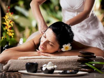 A woman is receiving a relaxing massage outdoors, adorned with a flower in her hair, surrounded by spa items and lush greenery.