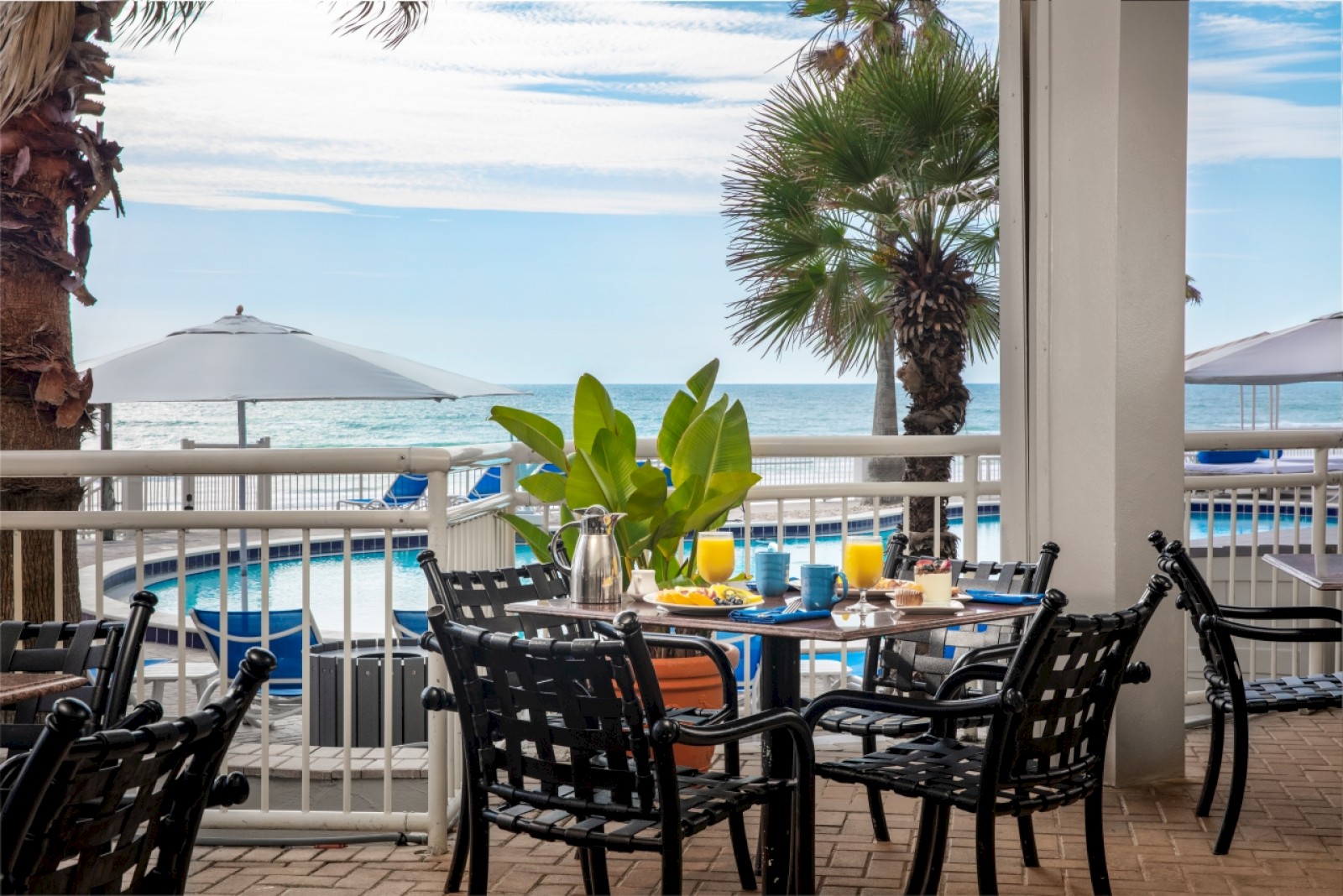 A seaside breakfast setting with a table of food, drinks, chairs, and umbrellas overlooking the ocean and palm trees.