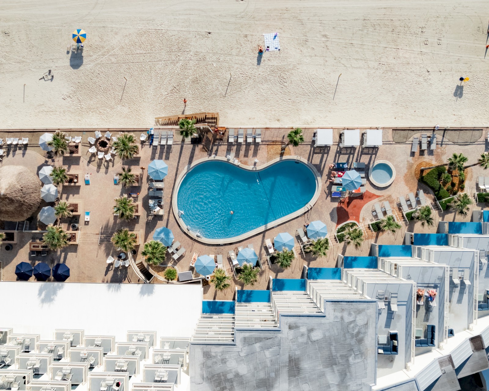 Vista aérea de un complejo turístico de playa que muestra una piscina en forma de corazón, tumbonas, sombrillas, palmeras y una playa de arena al fondo.