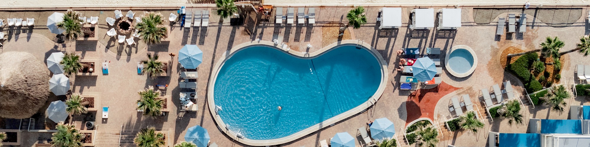 An aerial view of a beach resort showing a heart-shaped pool, lounge chairs, umbrellas, palm trees, and a sandy beach in the background.