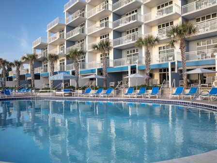 A pool with loungers in front of a hotel, clear skies above.