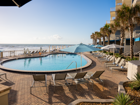 A serene beachside resort with a pool, sun loungers, umbrellas, and beachfront views of the ocean under clear skies.
