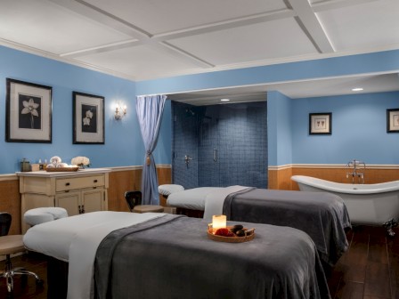 A luxurious spa room with two massage beds, a bathtub, and a shower, decorated with calming blue walls and framed floral artwork.