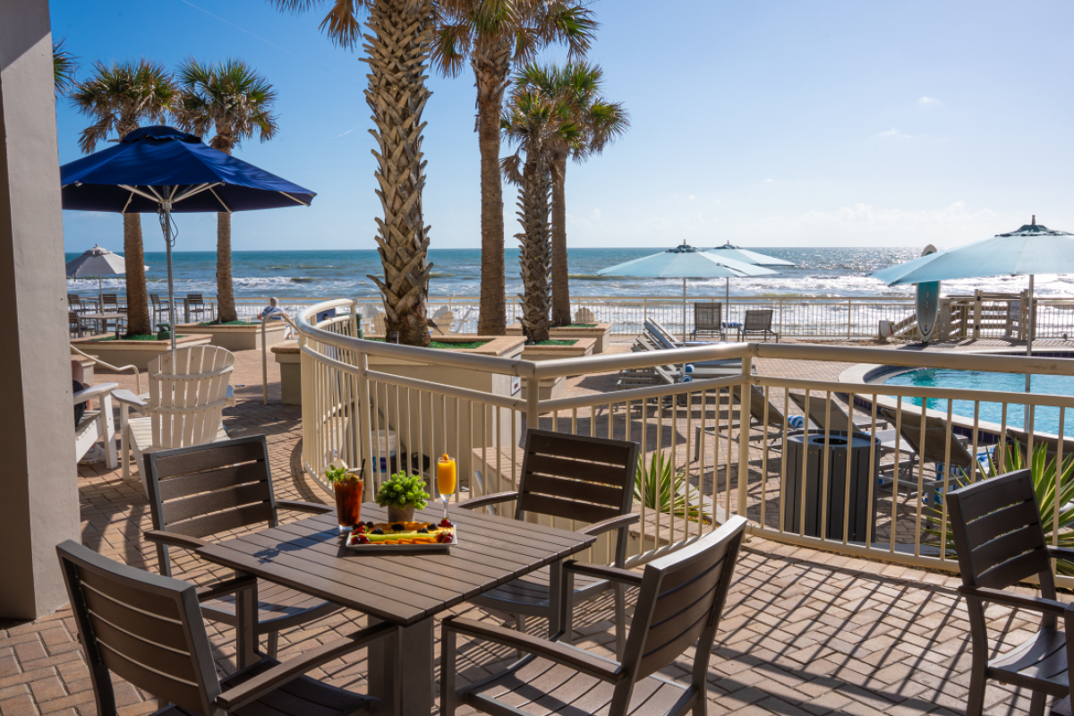 A seaside patio scene with a table set for breakfast, overlooking the ocean with palm trees, umbrellas, and lounge chairs.