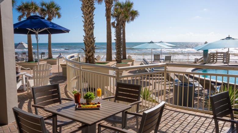 A seaside patio scene with a table set for breakfast, overlooking the ocean with palm trees, umbrellas, and lounge chairs.