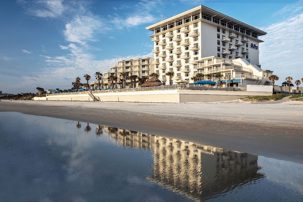 A seaside hotel with white walls is reflected in shallow water under a blue sky. Palm trees and beach with sand are visible in the background.