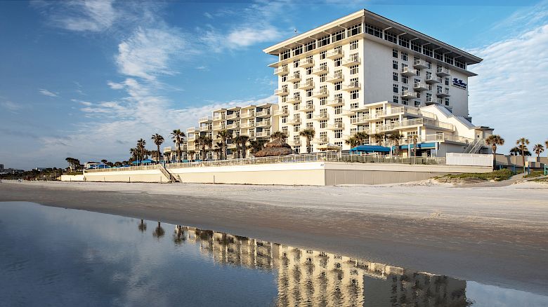 A seaside hotel with white walls is reflected in shallow water under a blue sky. Palm trees and beach with sand are visible in the background.