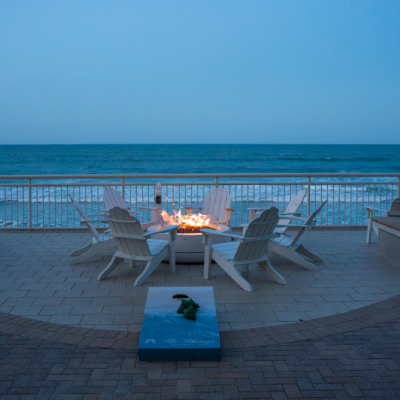 A cozy seaside patio with a fire pit, surrounded by chairs, overlooking the ocean at dusk.