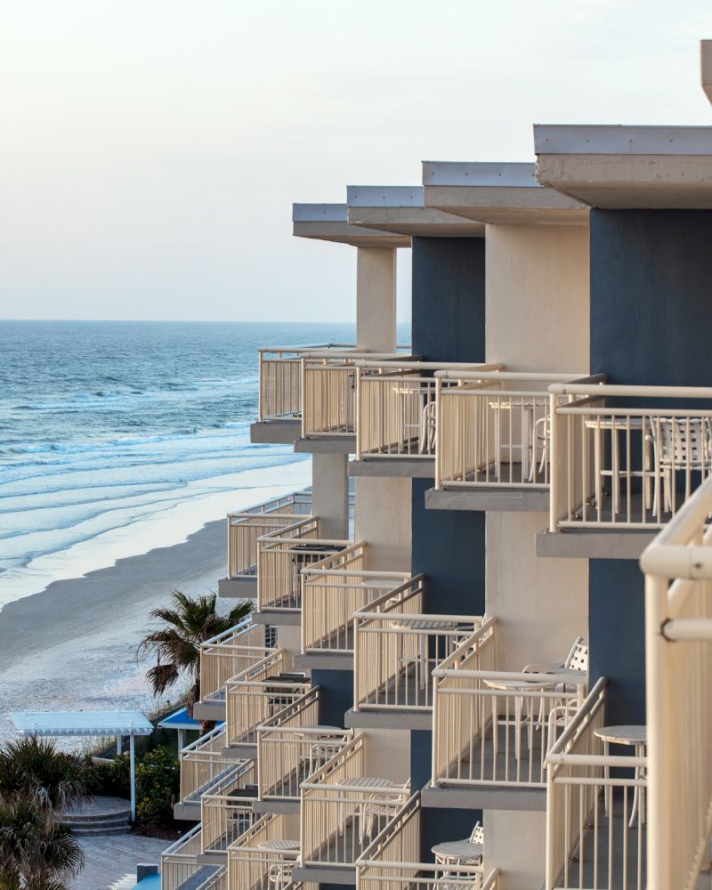 This image shows a beachside hotel with multiple balconies overlooking the ocean, palm trees along the shore, and calm waves on the sandy beach.