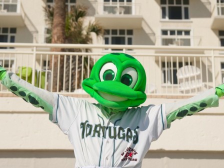 A person in a tortuga mascot costume wearing a baseball uniform with arms outstretched in front of a hotel building.