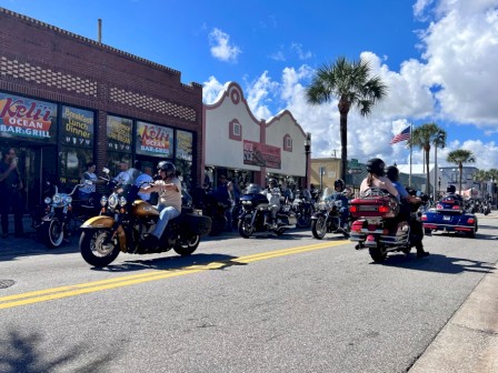 Motorcycles and people gather outside a diner on a sunny street with palm trees, American flags, and a bright blue sky.