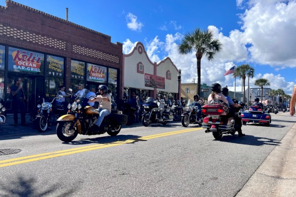 Motorcycles and people gather outside a diner on a sunny street with palm trees, American flags, and a bright blue sky.