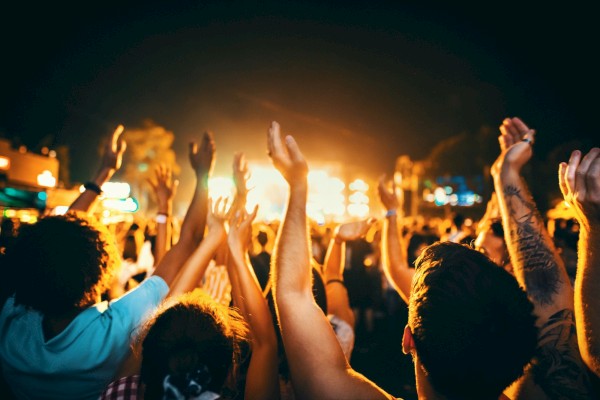 Crowd of people raising their hands at a concert or festival during nighttime with bright stage lights in the background.