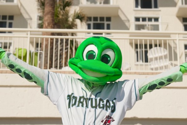 A person in a tortuga mascot costume wearing a baseball uniform with arms outstretched in front of a hotel building.