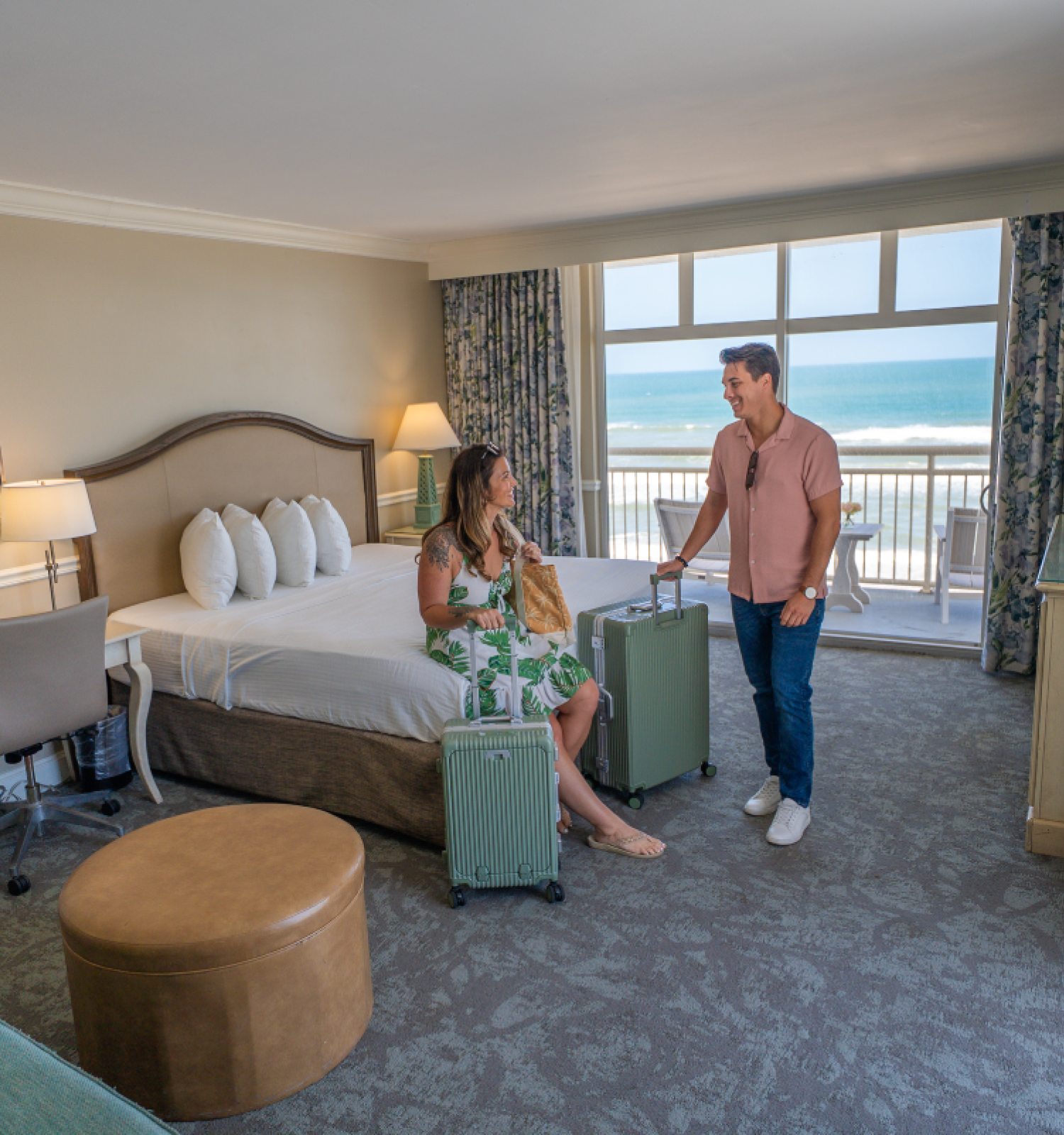 A woman sits on the bed with a woman standing, both with suitcases, inside a bright hotel room overlooking the ocean.