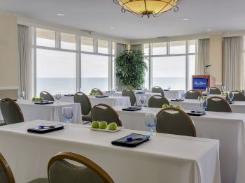 A conference room with tables and chairs arranged in rows, facing a podium. Each table has a notebook, green apples, and a glass of water ending the sentence.