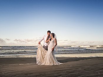 A couple in wedding attire stands on a beach with the ocean waves and a clear sky in the background, sharing a romantic moment, and posing.
