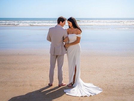 A couple in wedding attire shares a romantic moment on the beach under a bright blue sky.