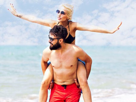 A man in red shorts carries a woman in black swimwear on his back along a beach. The woman has her arms outstretched and both are smiling.