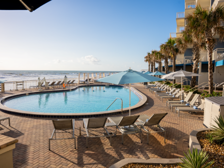 A swimming pool overlooking the ocean with lounge chairs and umbrellas, set in a seaside hotel with palm trees on a sunny day.