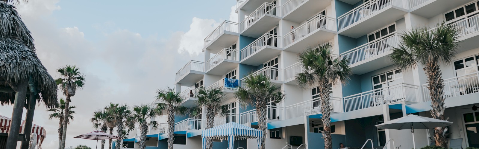 A resort-style building with balconies and palm trees is reflected in a large swimming pool under a clear sky.