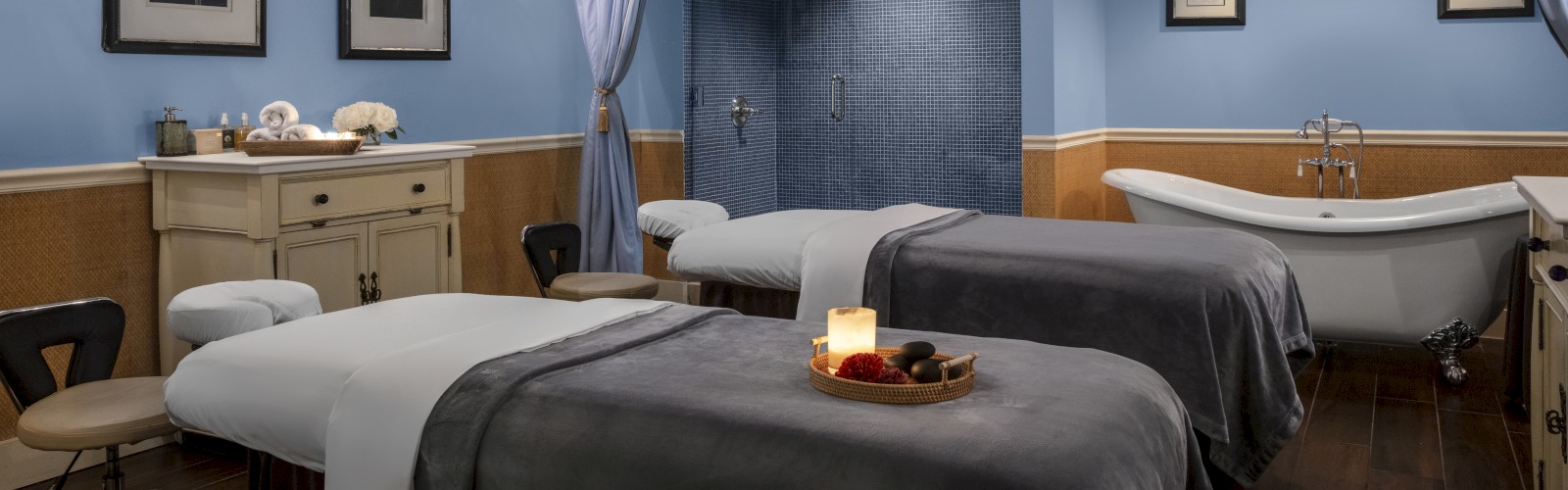 A relaxing spa room with two massage tables, a bathtub, and soft lighting. The decor is calming, with light blue walls and framed art.