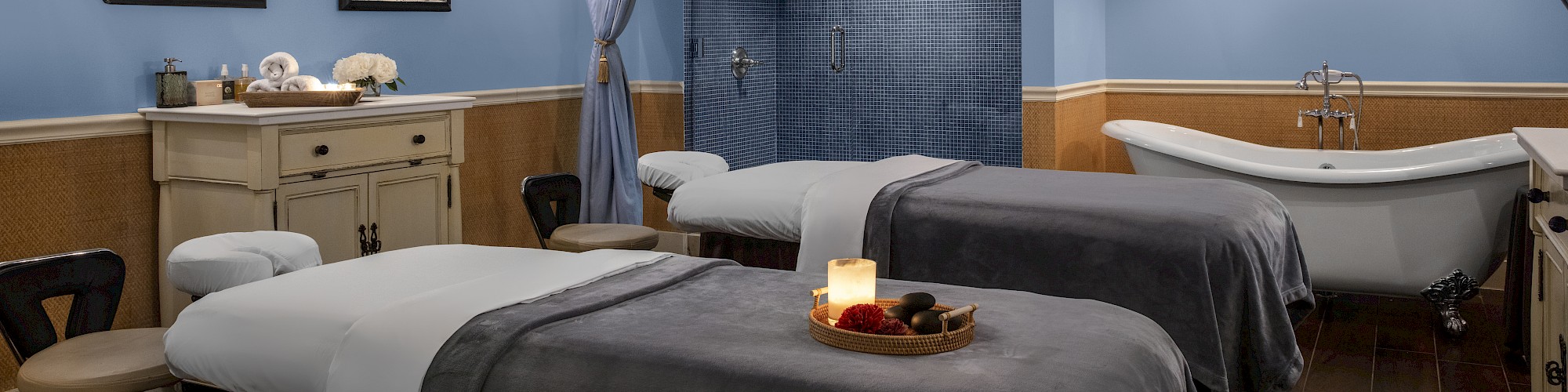 A relaxing spa room with two massage tables, a bathtub, and soft lighting. The decor is calming, with light blue walls and framed art.