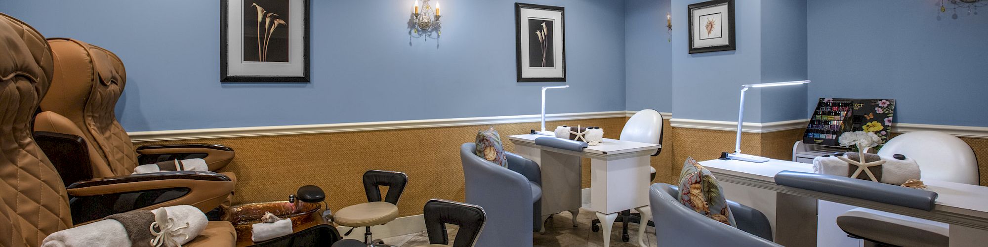 This image shows an interior of a nail salon with pedicure chairs, manicure stations, blue walls, framed artwork, and ceiling lights.