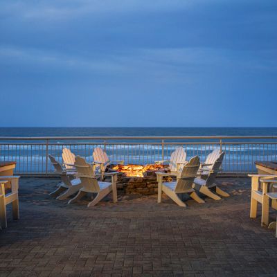 A seaside view with white Adirondack chairs arranged around a fire pit on a brick patio, flanked by palm trees under a twilight sky, ending the sentence.