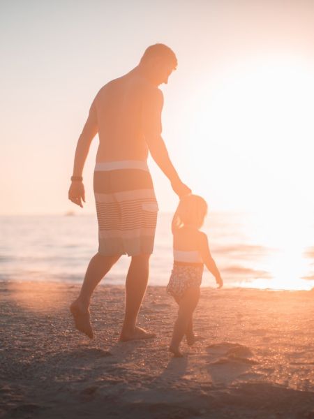 A person and a child walking hand in hand on a beach at sunset, with the ocean in the background.
