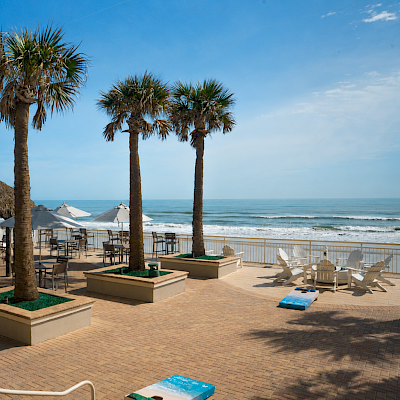 A relaxing beachside scene with palm trees, thatched hut, patio furniture, and ocean waves on a sunny day.