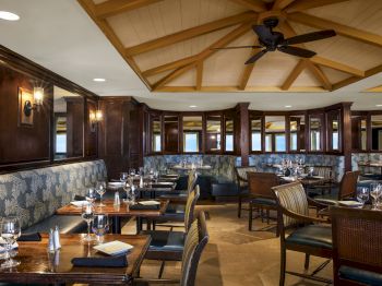 This image shows a clean, well-lit restaurant with wooden tables, cushioned seating, and place settings, featuring a ceiling fan and patterned walls.