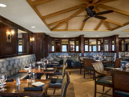 This image shows a clean, well-lit restaurant with wooden tables, cushioned seating, and place settings, featuring a ceiling fan and patterned walls.