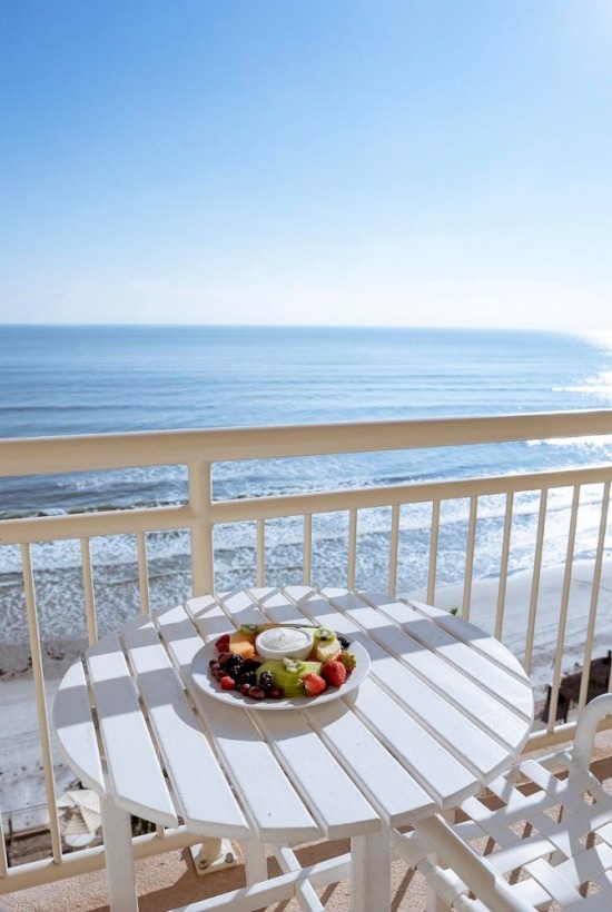 A balcony overlooks the beach with a table of fresh fruit and a scenic ocean view under bright sunlight.