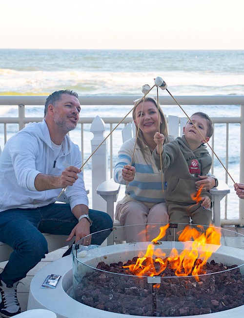 Four people, two adults, one child, and a teenager, sit around a fire pit on a porch by the ocean, roasting marshmallows and enjoying each other's company.