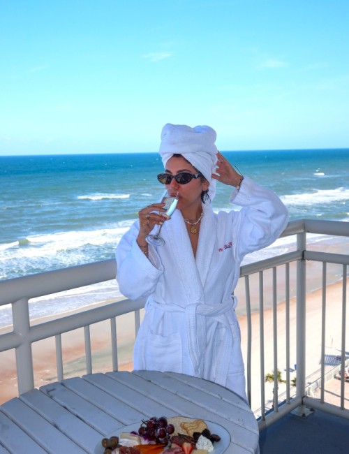 A woman in a bathrobe and towel on her head enjoys a drink, with a cheese platter and sea view on a balcony.