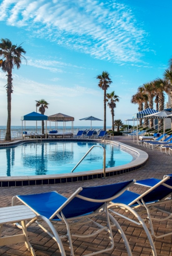 A sunny poolside scene at a beach hotel, with empty lounge chairs, umbrellas, palm trees, and a view of the ocean.