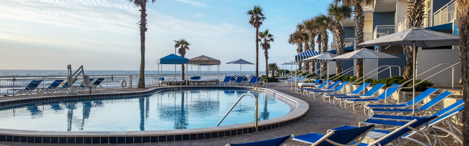 A seaside resort with a pool, surrounded by blue lounge chairs, beach umbrellas, palm trees, and a view of the ocean under a partly cloudy sky.