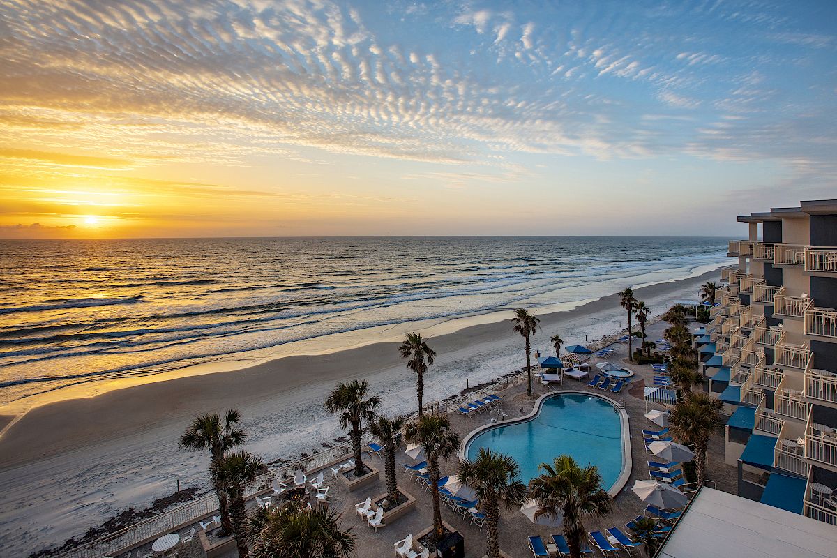 The image shows a beachside view at sunset, featuring a swimming pool, palm trees, lounge chairs, and a multi-story hotel/building on the right.