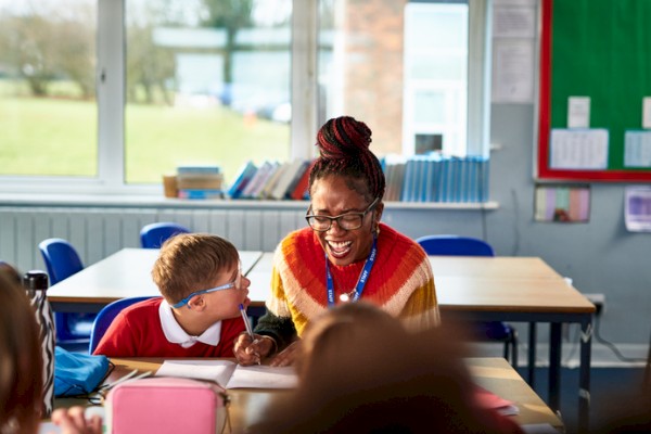A joyful teacher or volunteer interacting with a young student in a classroom setting with books and learning materials.